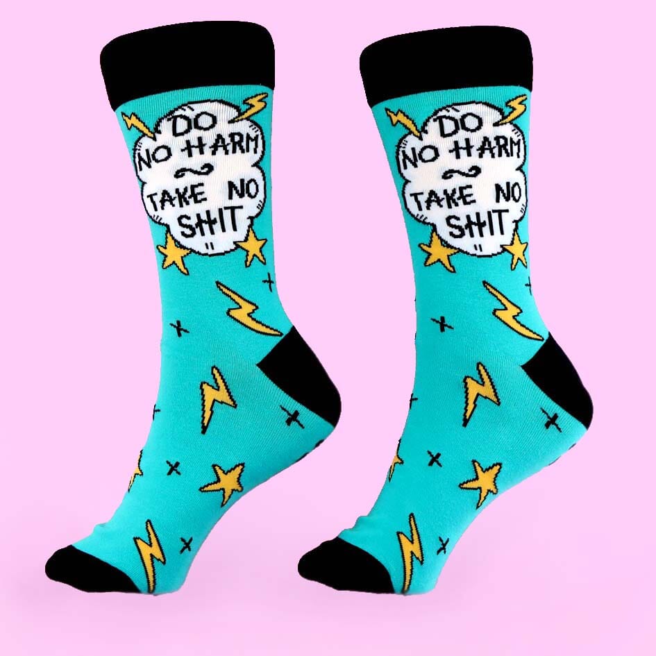 A pair of socks standing against a pink background. The socks are teal and black and read Do No Harm Take No Shit.