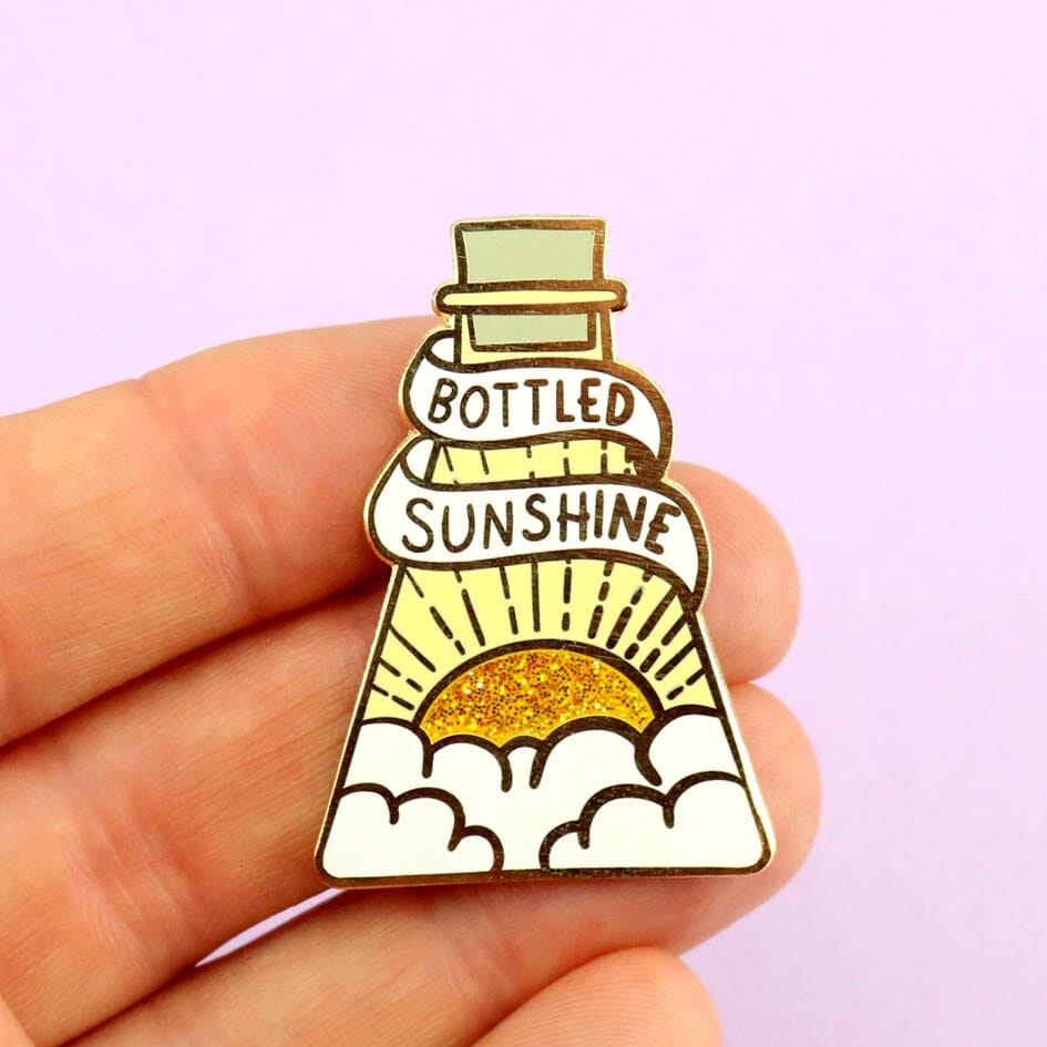 Pin: A hard enamel lapel pin being held in a hand. The pin is in the profile of a beaker with a yellow glitter sun. The pin says Bottled Sunshine.