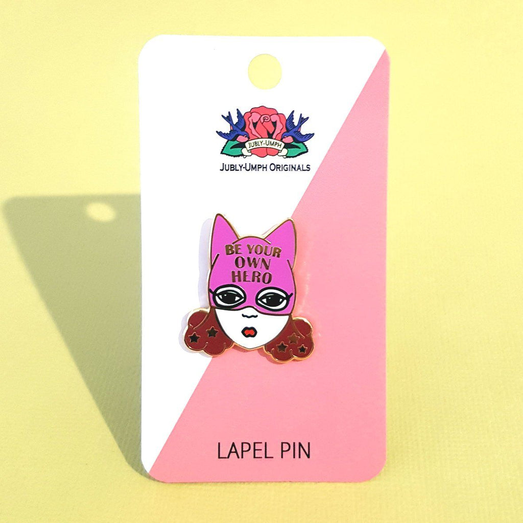 A hard enamel lapel pin on Jubly-Umph cardstock. The pin is a white woman with brown hair and a superhero hat that says Be Your Own Hero.