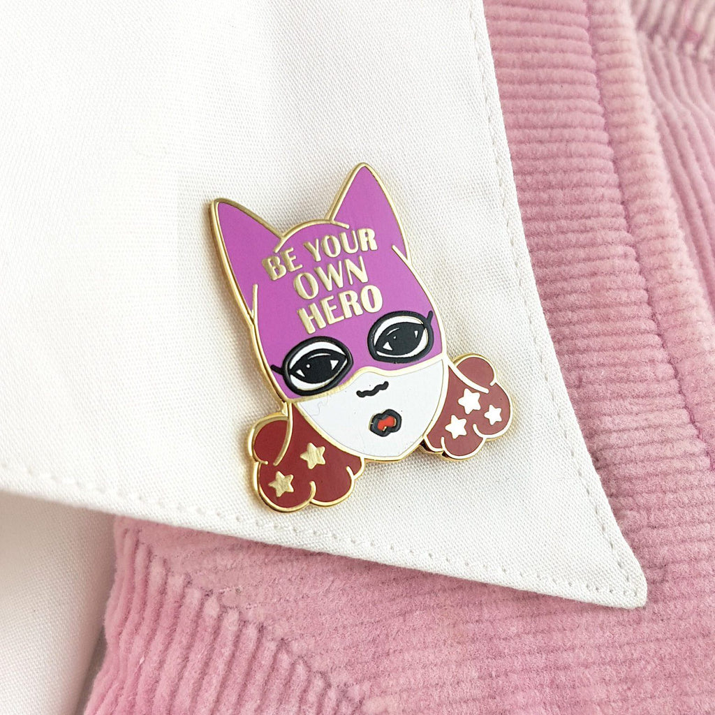 A hard enamel lapel pin on a white shirt collar. The pin is a white woman with brown hair and a superhero hat that says Be Your Own Hero.