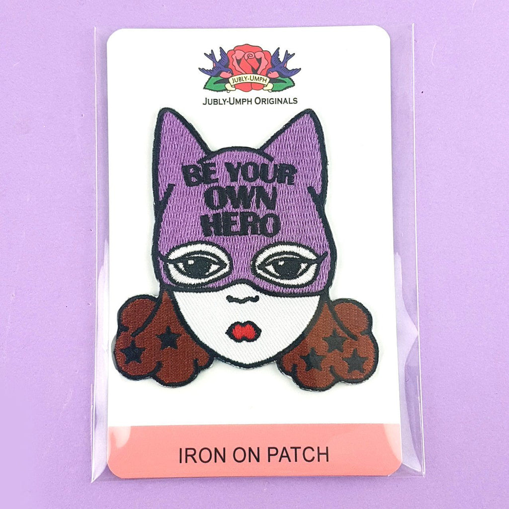 An iron on embroidered patch on Jubly-Umph cardstock against a purple background. The patch is a white woman with brown hair and a superhero hat that says Be Your Own Hero.