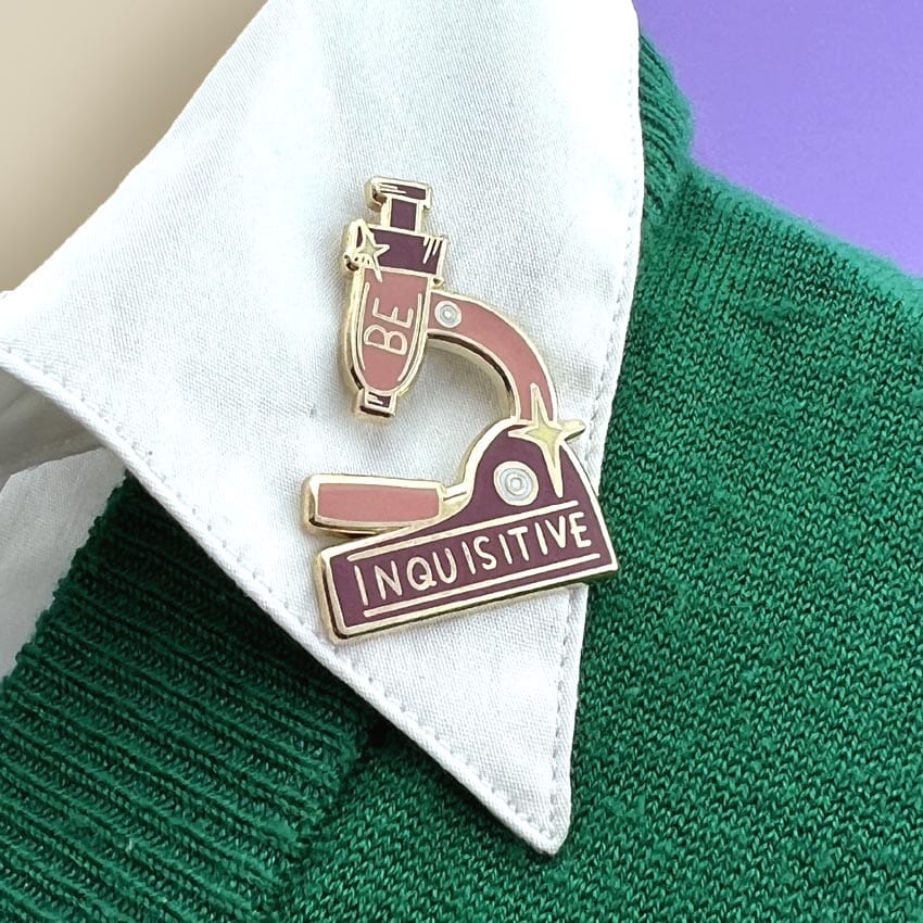 A hard enamel lapel pin being worn on a white collar. The pin is in the shape of a microscope and reads Be Inquisitive.