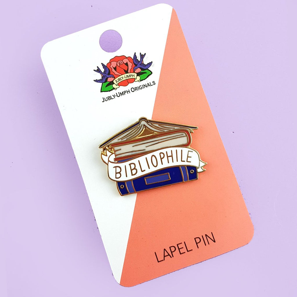 A hard enamel lapel pin on a purple background. The pin says Bibliophile in the middle of a stack of blue books.