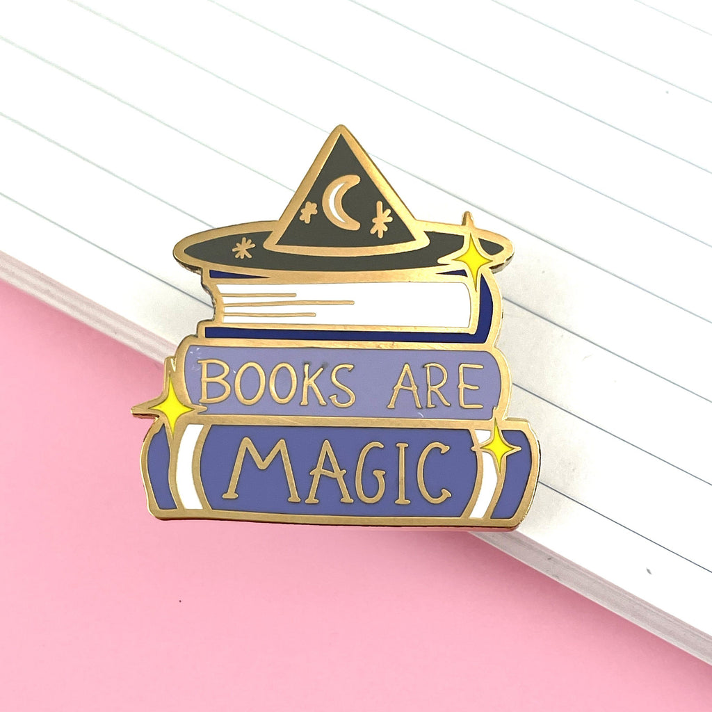A hard enamel lapel pin on a lined paper background. The pin is purple and white and says Books are Magic with books and a witch’s hat.  