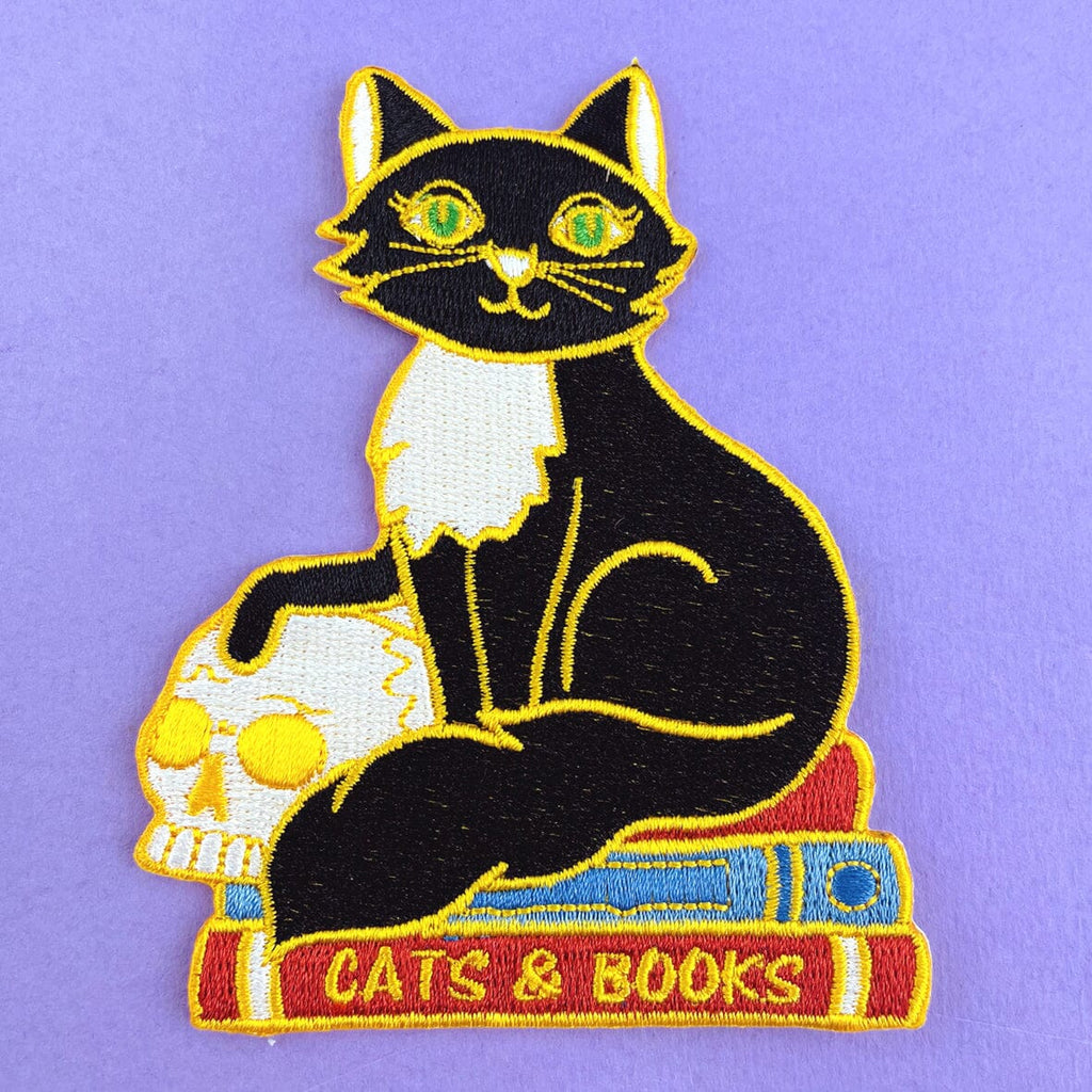 An iron on embroidered patch against a purple background. The patch is a black and white cat with a scull sitting on books. The patch reads Cat’s and Books.