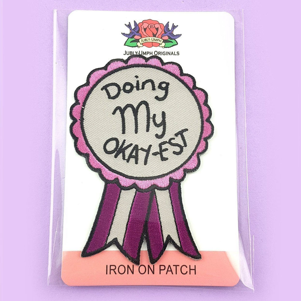 An iron on embroidered patch on Jubly-Umph cardstock. The patch is in the shape of an award ribbon. The ribbon is pink and grey and reads Doing my Okay-est.