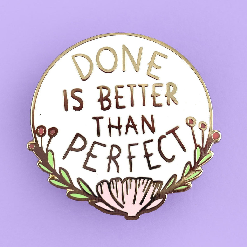 A hard enamel lapel pin on a light purple background. The pin is white with gold lettering and reads Done Is Better Thank Perfect.