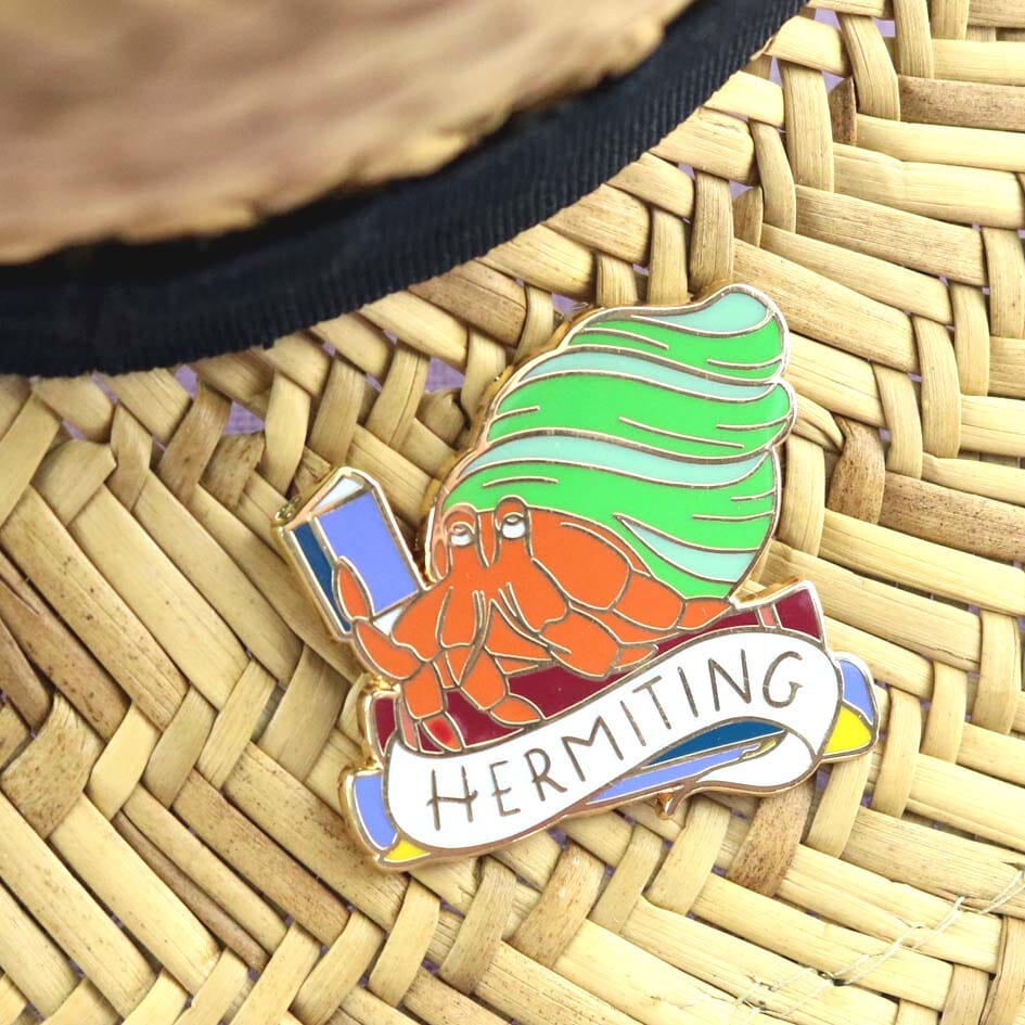 A hard enamel lapel pin on a straw hat background. The pin is in the shape of a Hermit Crab reading a book. The pin says Hermit Crab.