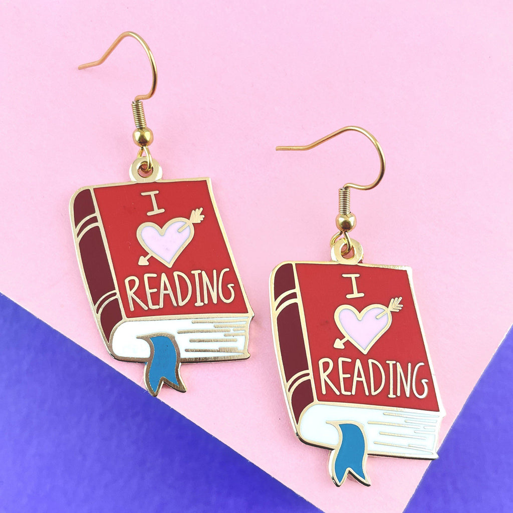 A pair of earrings displayed on a pink background. The earrings are in the shape of a red book. The earrings read I (heart image) reading.