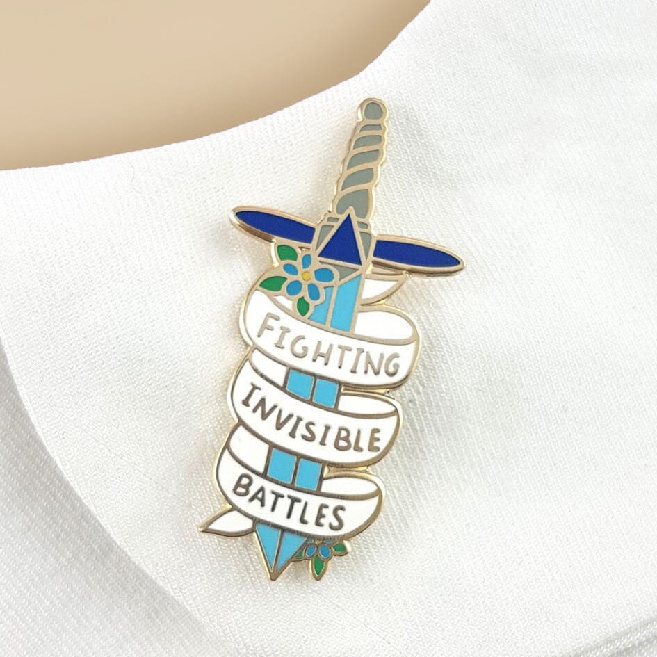 A hard enamel pin on a white lapel. The pin is in the shape of a dagger and reads Fighting Invisible Battles.