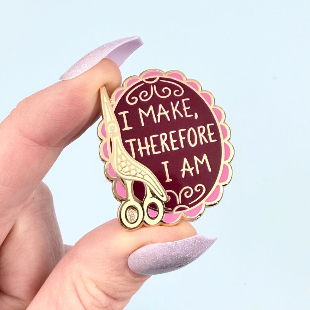 Pin with scissors and text 'I make, therefore I am' held by a hand against a light blue background