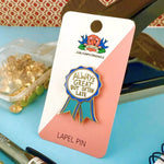 Lapel pin with text "Always great but often late" on a card with 'Jubly-Umph Originals' branding, placed on a blue surface with office supplies.