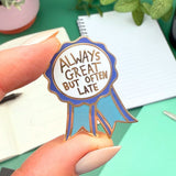 Enamel pin with 'Always great but often late' text held by a hand on a green surface.