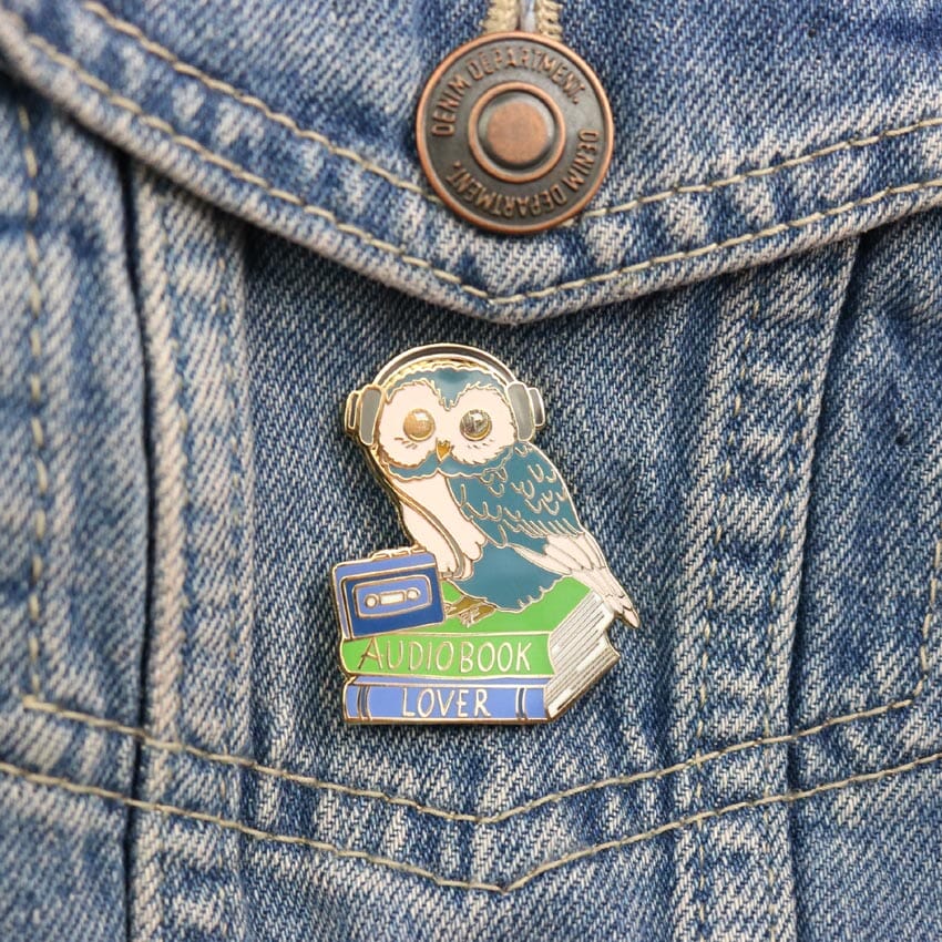 A hard enamel lapel pin against a denim jacket. The pin is in the shape of an owl sitting on a stack of books wearing headphones. The pin reads Audiobook Lover.