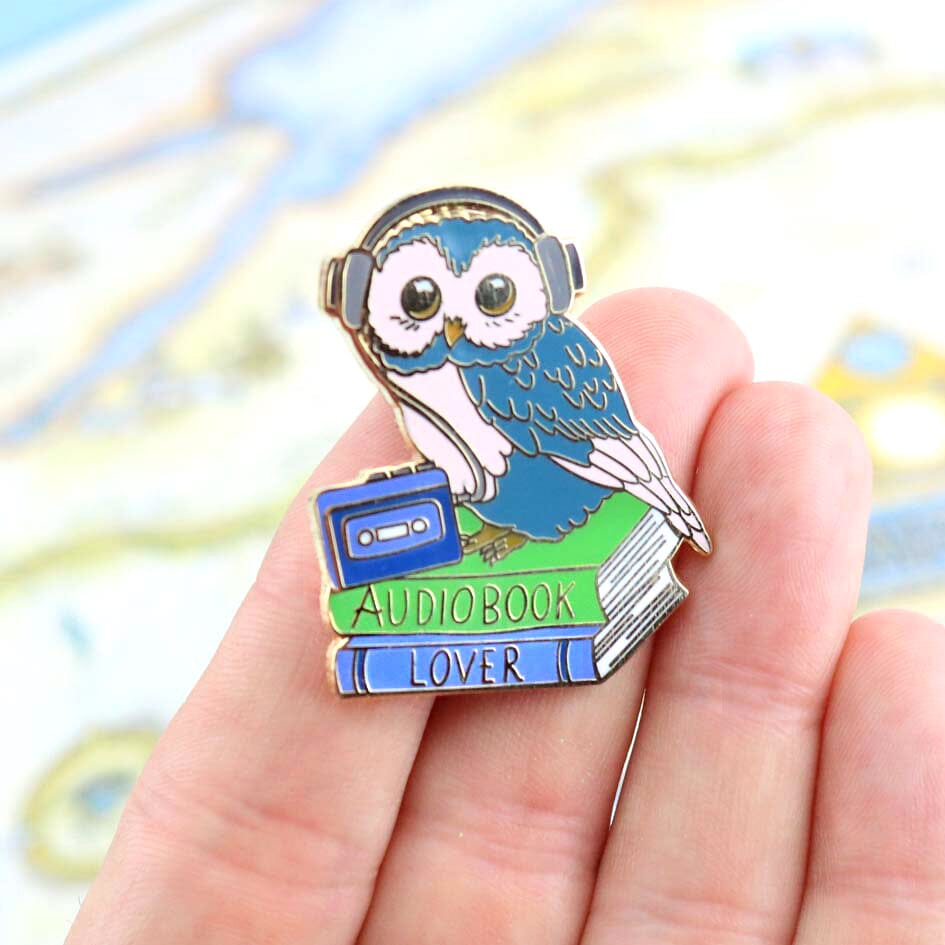 A hard enamel lapel pin being held in a hand. The pin is in the shape of an owl sitting on a stack of books wearing headphones. The pin reads Audiobook Lover.