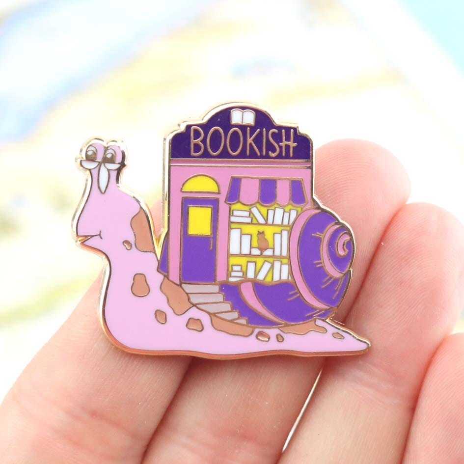 A hard enamel lapel pin being held in a hand. The pin says say Bookish. The pin design is a snail with a bookshop on its back.