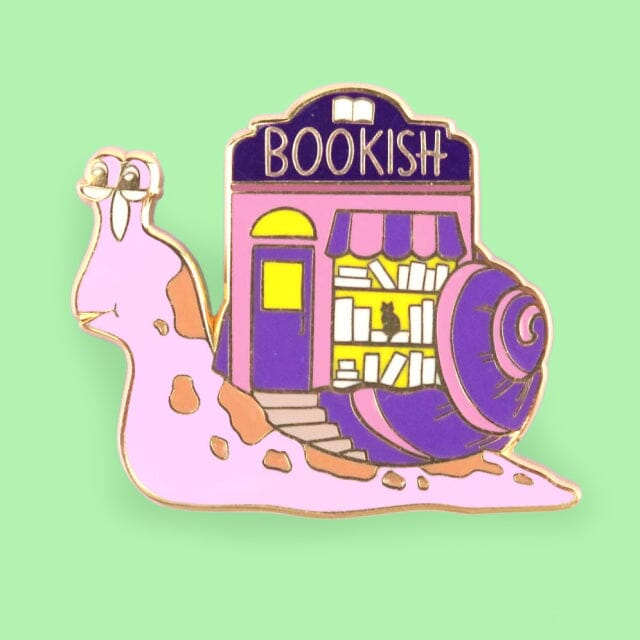 A hard enamel lapel pin on a green background. The pin says say Bookish. The pin design is a snail with a bookshop on its back.