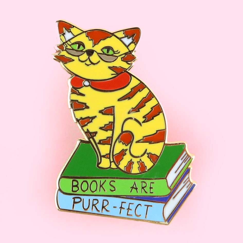 A hard enamel lapel pin on a pink background. The pin says Books Are Purr-fect with a cat sitting on books. 