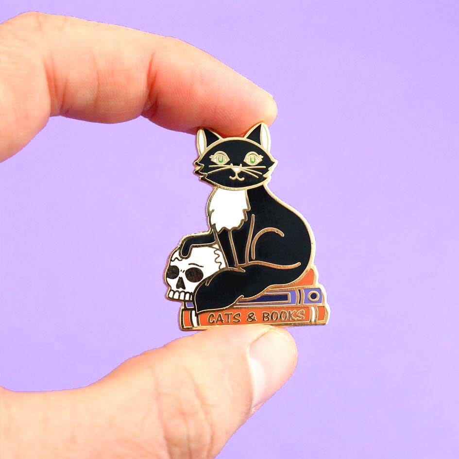 A hard enamel lapel pin being held in a hand. The pin is in the shape of a black and white cat with a scull, sitting on books. The pin reads Cat’s and Books.