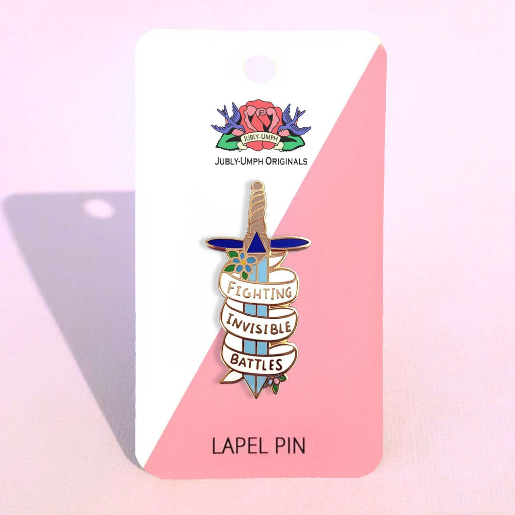 A hard enamel pin on a Jubly-Umph backing card. The pin is in the shape of a dagger and reads Fighting Invisible Battles.