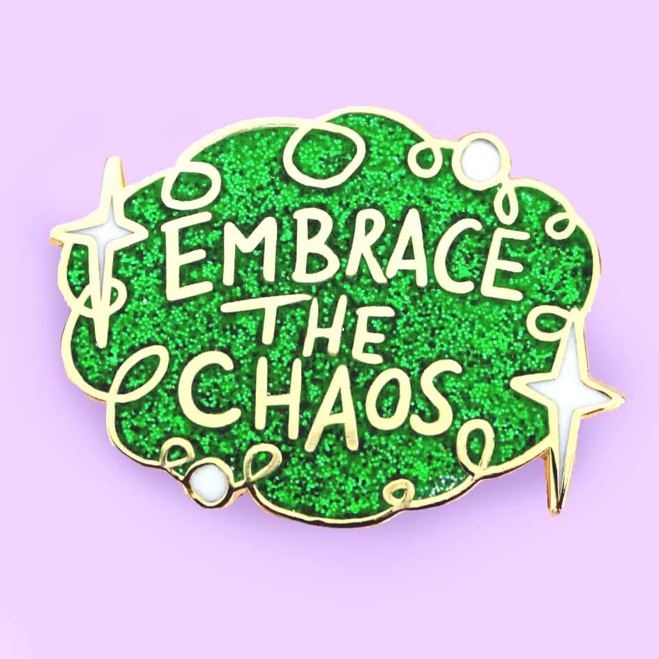 A hard enamel lapel pin on a purple background. The pin is green glitter with white stars and reads Embrace the Chaos.