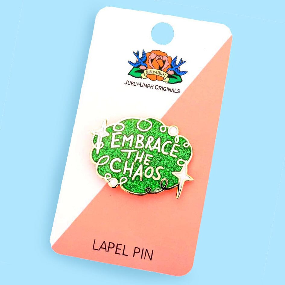 A hard enamel pin on Jubly-Umph card stock with a blue background. The pin is green glitter with white stars and reads Embrace the Chaos.