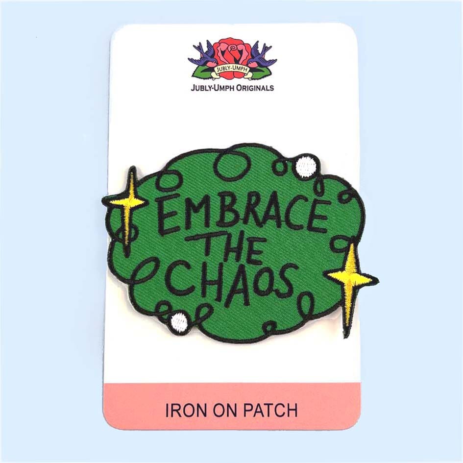 An iron on embroidered patch on Jubly-Umph cardstock against a blue background. The patch is green with yellow stars and reads Embrace The Chaos.