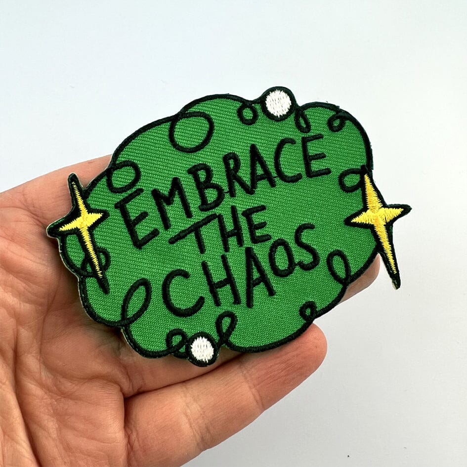 An iron on embroidered patch being held in a hand against a blue background. The patch is green with yellow stars and reads Embrace The Chaos.