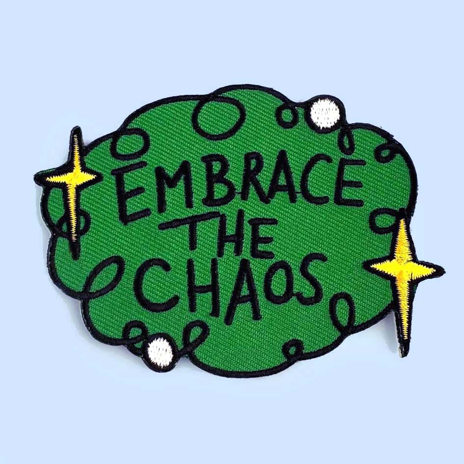 An iron on embroidered patch against a blue background. The patch is green with yellow stars and reads Embrace The Chaos.