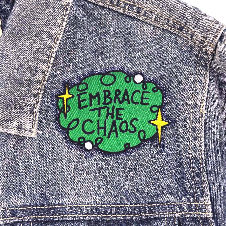 An iron on embroidered patch on a denim jacket. The patch is green with yellow stars and reads Embrace The Chaos.