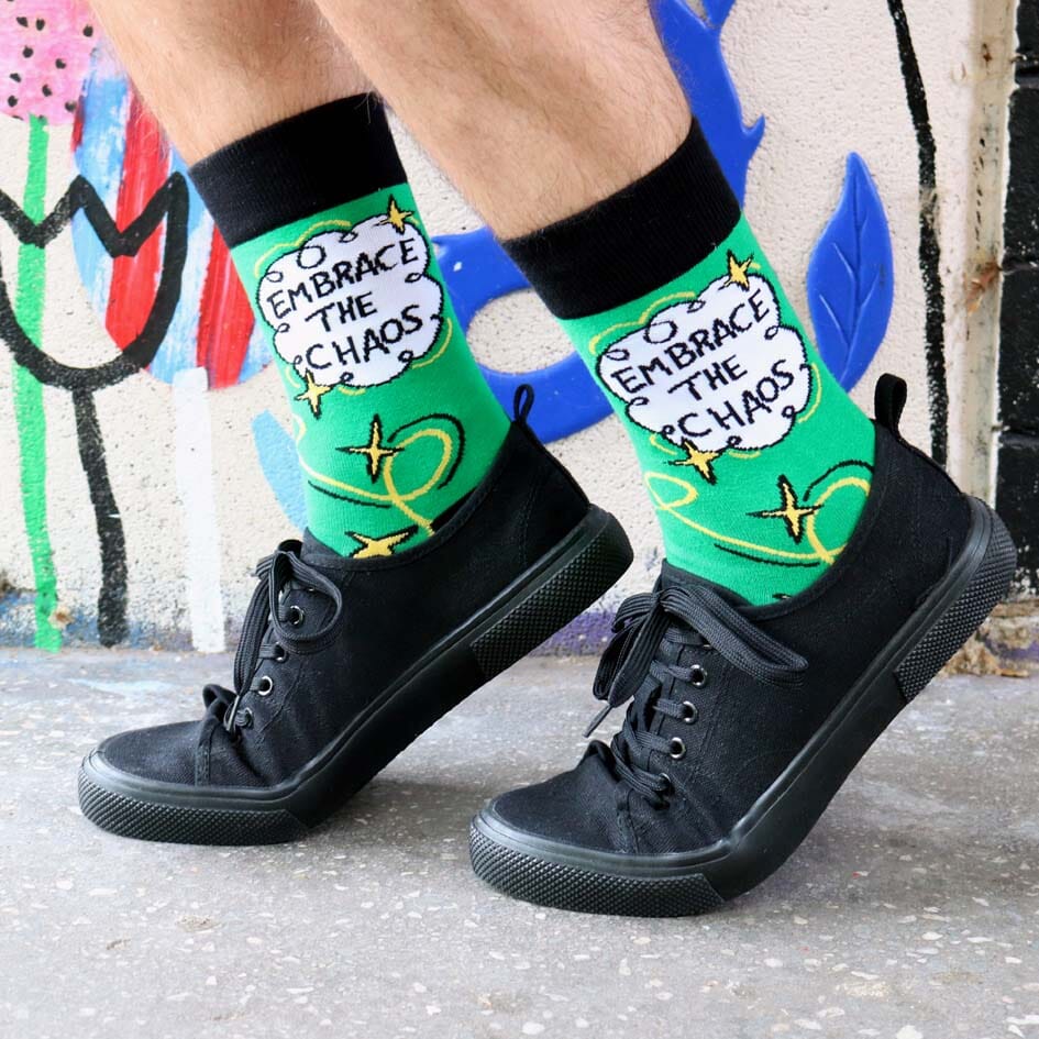 A pair of socks being worn with black shoes standing against a graffiti wall. The socks are green and gold and read Embrace The Chaos. 