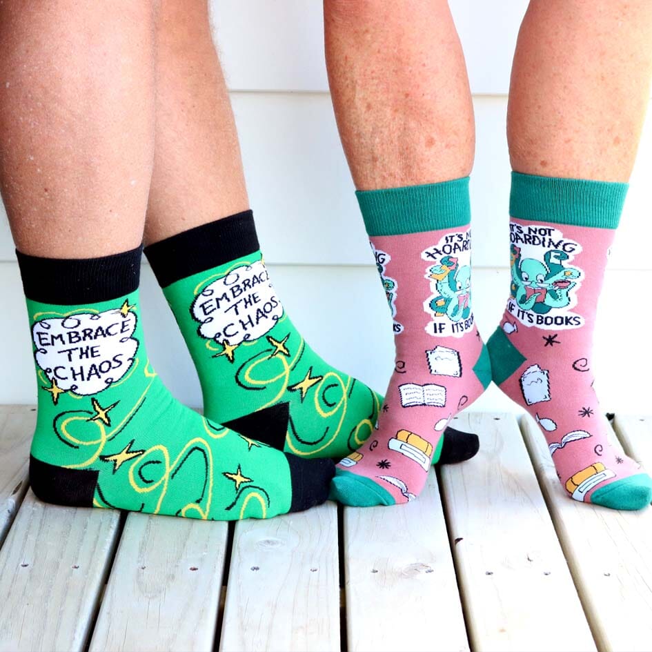 Two pairs of socks being worn. The firdt pair of socks socks are green and gold and read Embrace The Chaos. The second pair reads It's not hoarding if it's books.