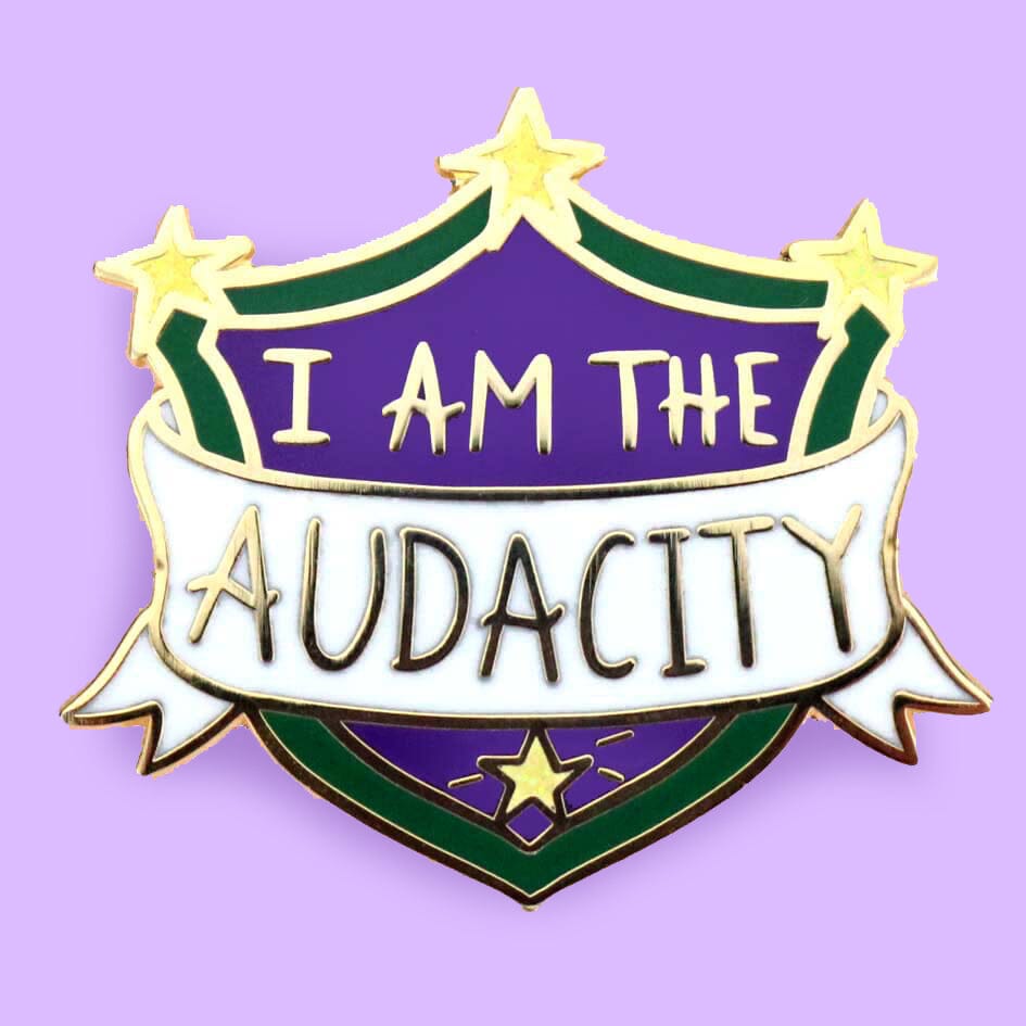A hard enamel lapel pin on a purple background. The pin is in the shape of a shield. The lapel pin reads I am the Audacity.