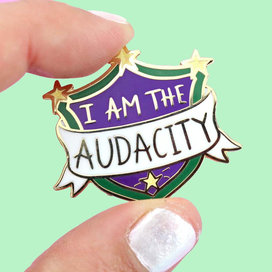 A hard enamel lapel pin being held in a hand. The pin is in the shape of a shield. The lapel pin reads I am the Audacity.