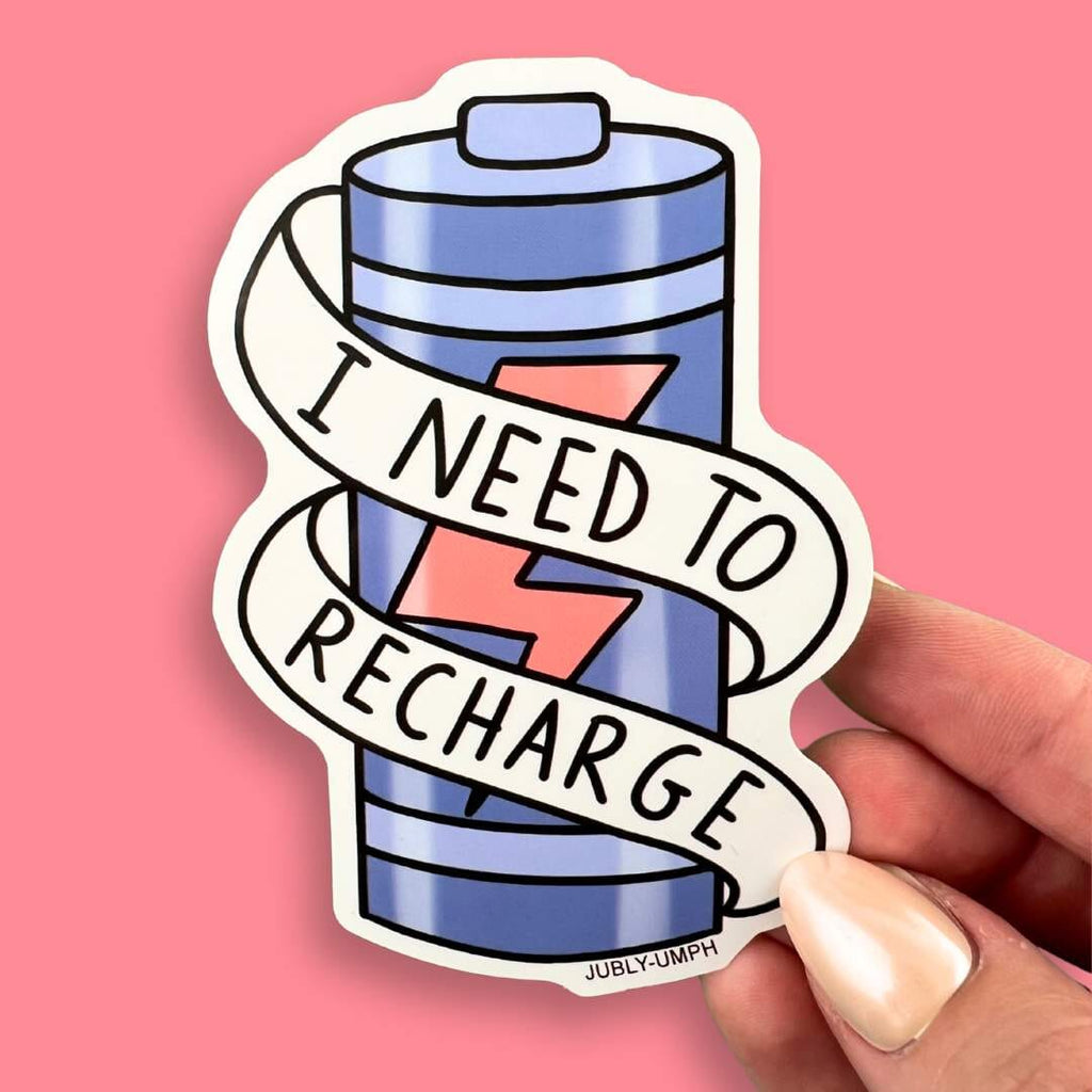 The vinyl sticker is in the shape of a battery being held in the hand against a pink background. The sticker reads I Need To Recharge.
