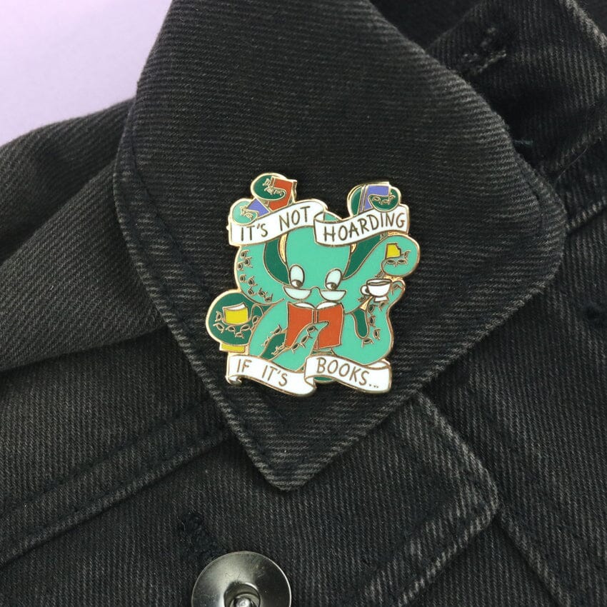 A hard enamel lapel pin on black jean jacket. The pin says It’s Not Hoarding If It's Books in the form of an octopus.