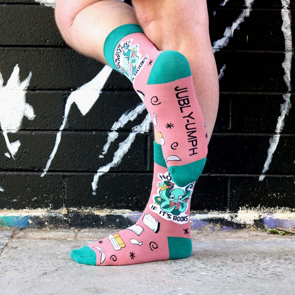 A pair of socks are being worn by a model. The bottom of one foot is being shown. It reads Jubly-Umph. The socks are green and light brown with a light green octopus. The socks read It's Not Hoarding If It's Books.