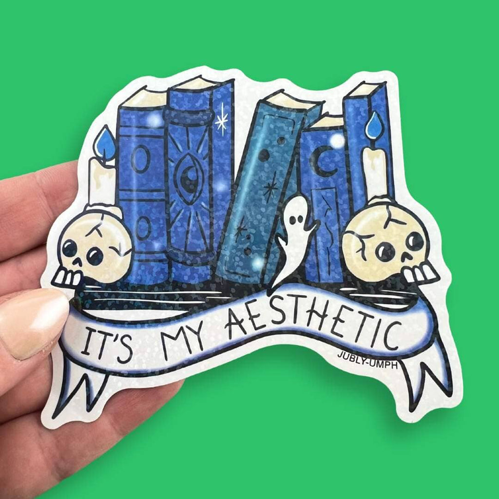 The vinyl sticker is in the shape of books on a shelf with skulls and candles. It is being held in a hand on a green background. The sticker is blue with glitter and reads It's My Aesthetic.