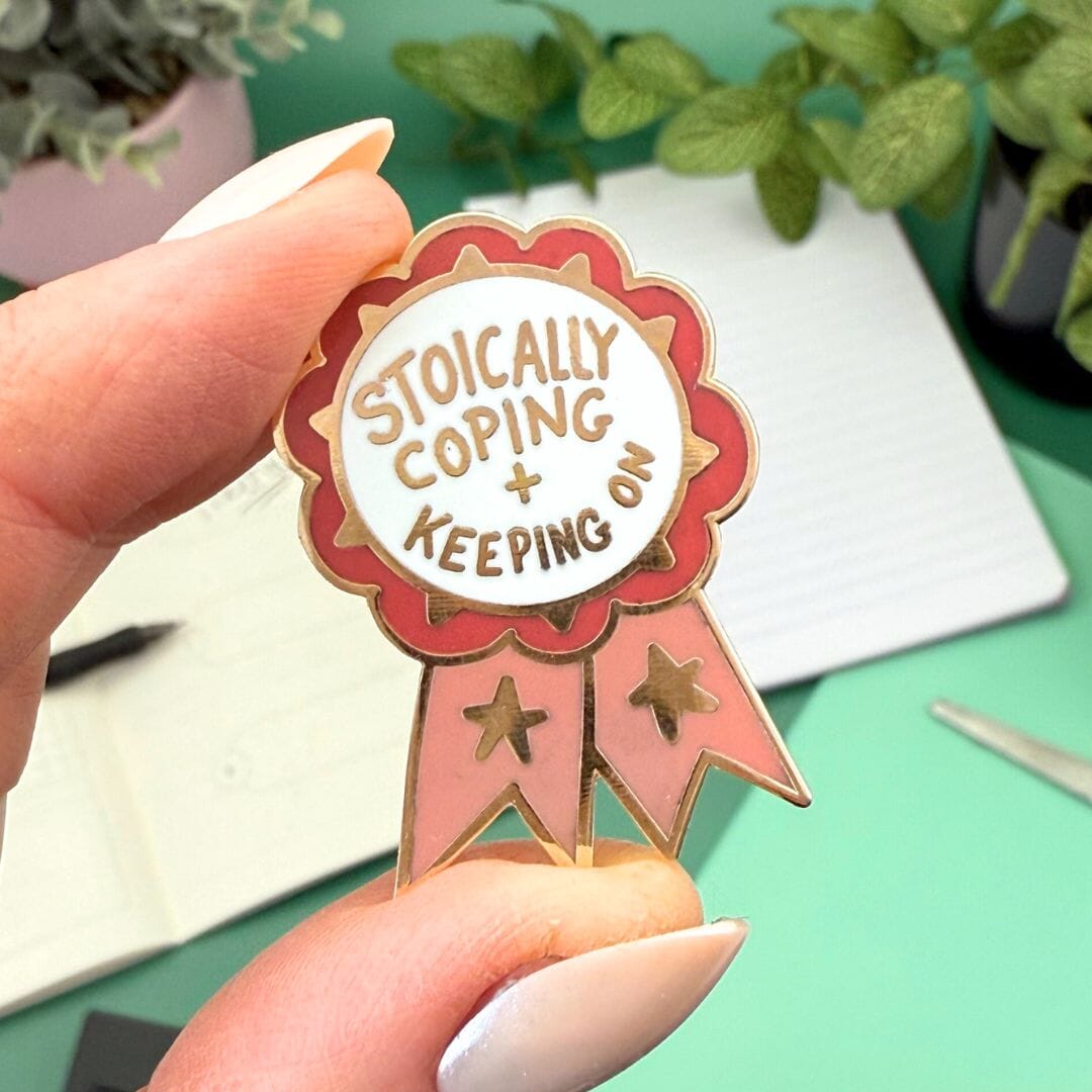 Enamel pin with 'Stoically Coping & Keeping' text held by a hand against a green background with plants.