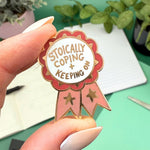 Enamel pin with 'Stoically Coping & Keeping' text held by a hand against a green background with plants.