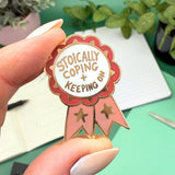Enamel pin with 'Stoically Coping & Keeping' text held by a hand against a green background with plants.