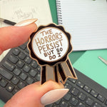 Pin with 'The Horrors Persist But So Do I' text held by a hand on a desk.