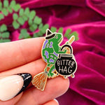 Bitter Hag pin being held with plant and velvet red background