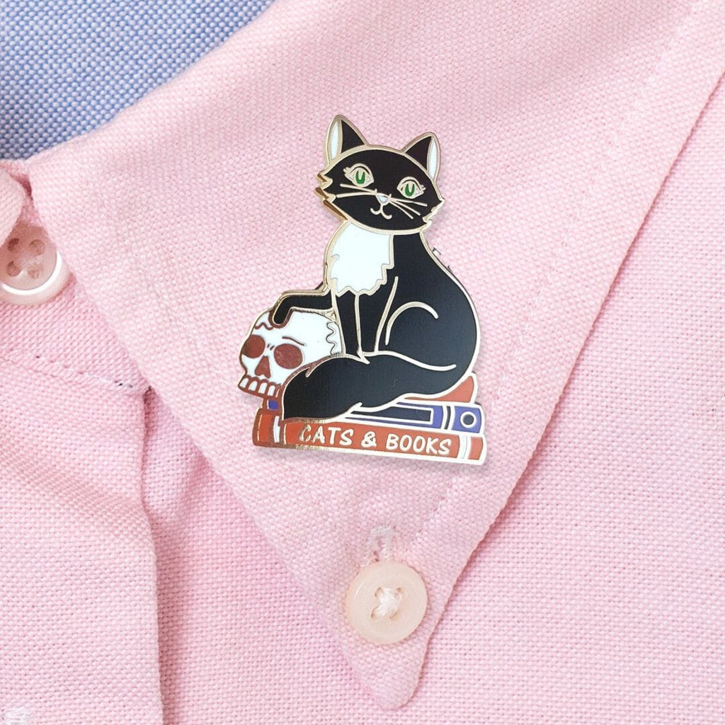 A hard enamel lapel pin on a pink shirt. The pin is in the shape of a black and white cat with a scull, sitting on books. The pin reads Cat’s and Books.