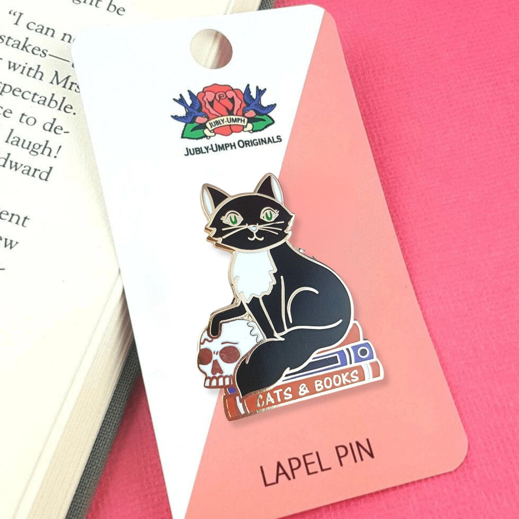 A hard enamel lapel pin on Jubly-Umph cardstock. The pin is in the shape of a black and white cat with a scull, sitting on books. The pin reads Cat’s and Books.