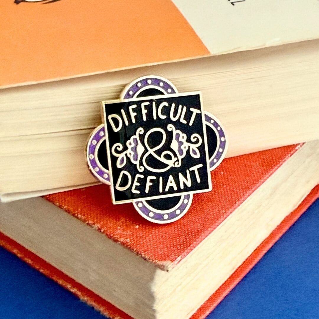 Enamel pin with 'Difficult & Defiant' text on a stack of books