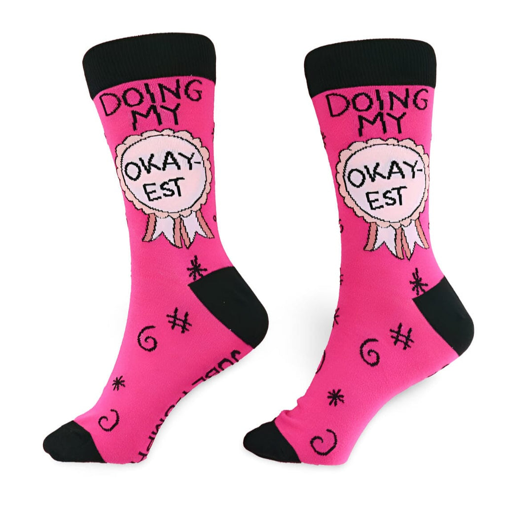 A pair of socks on a white background. The socks are pink and black and read Doing My Okay-est.