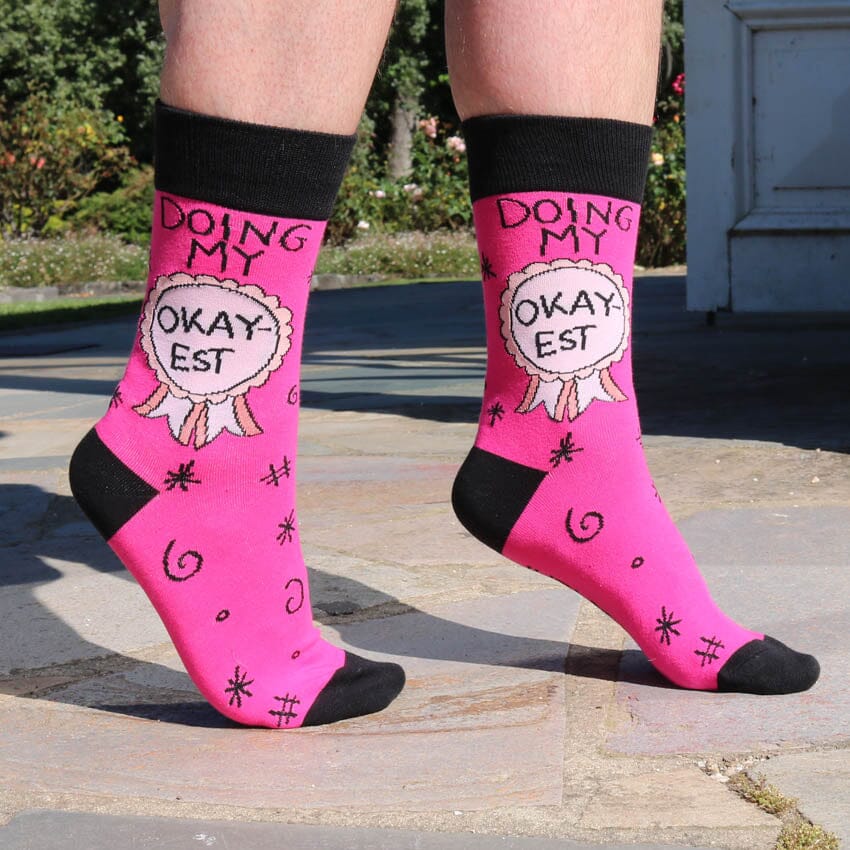 A pair of socks being modelled on tip toes. The socks are pink and black and read Doing My Okay-est.