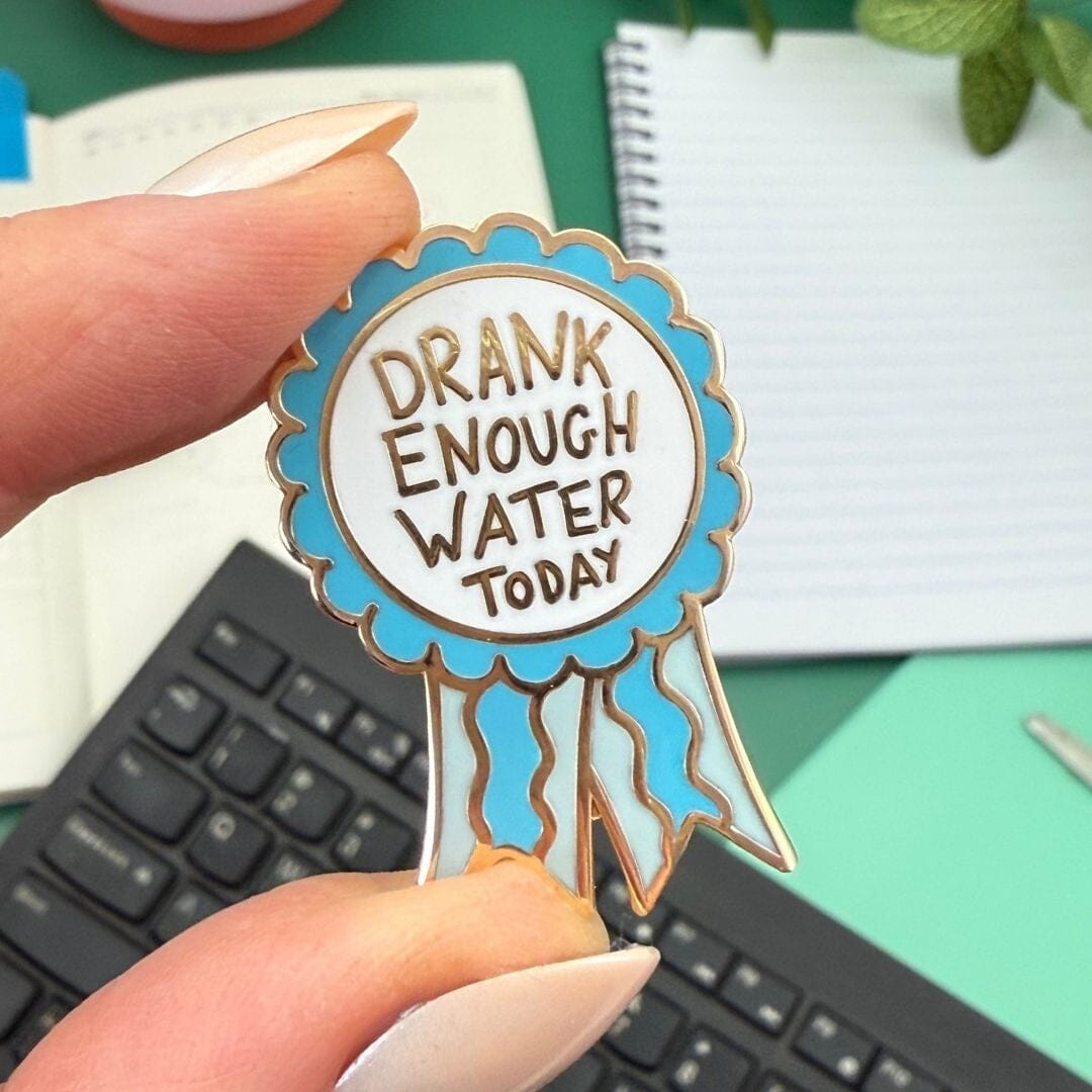 Pin with 'Drank Enough Water Today' text held by a hand on a desk.
