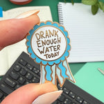 Pin with 'Drank Enough Water Today' text held by a hand on a desk.